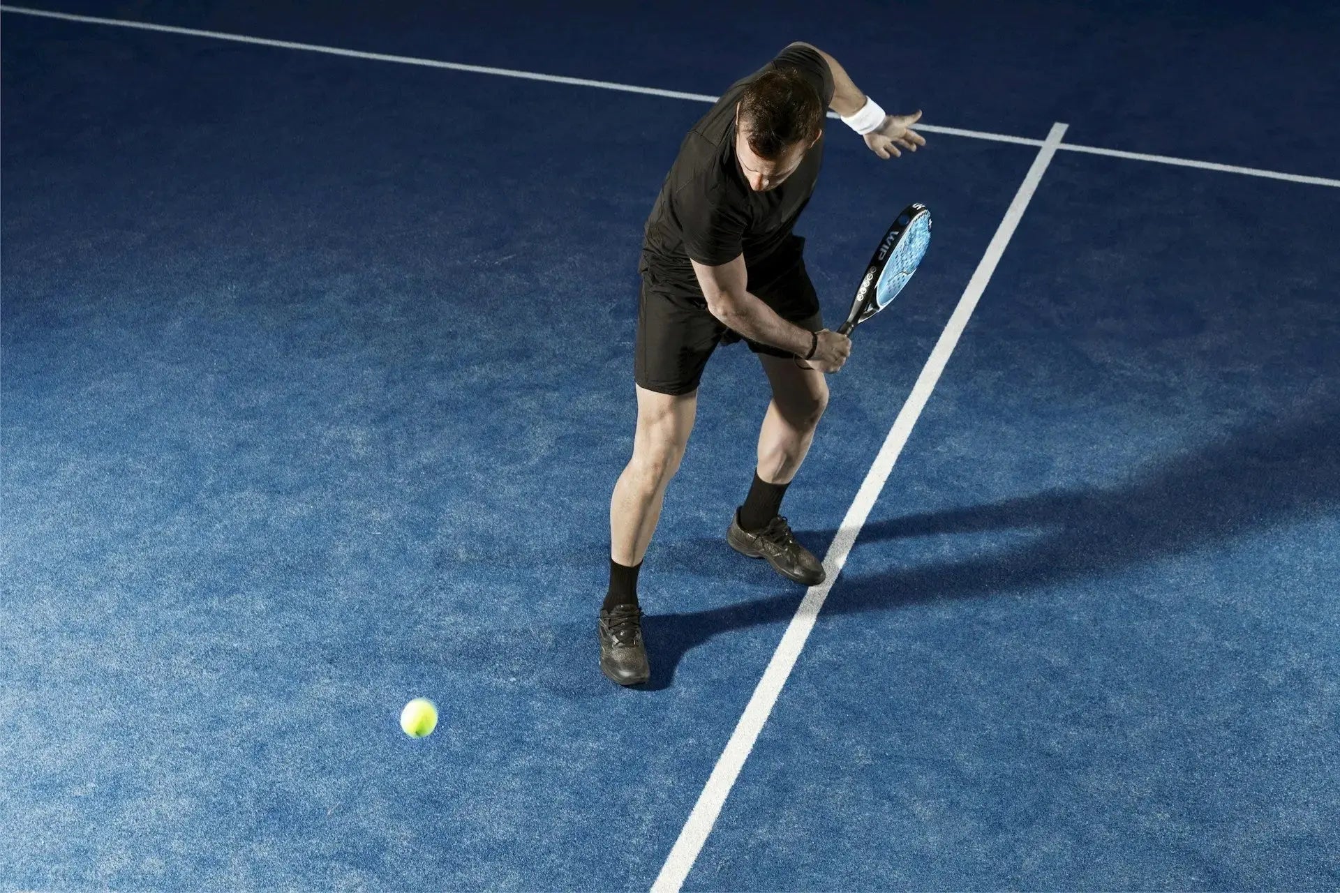 Homme en tenue noire jouant au padel et frappant une balle basse sur un terrain bleu